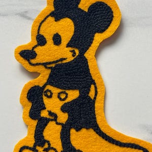 May include: A yellow felt patch featuring black embroidered outlines of a cartoon mouse character. The design shows a larger mouse figure standing behind a smaller mouse figure, both rendered in a simple, bold, and graphic embroidery style.