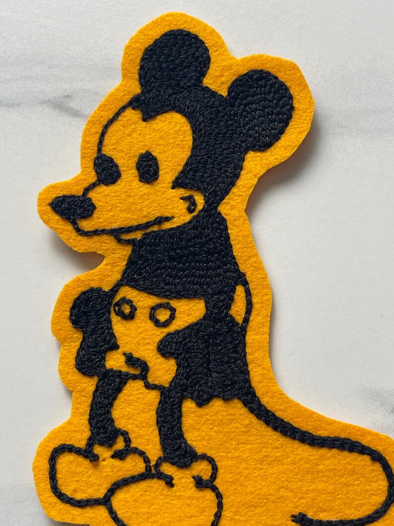 May include: A yellow felt patch featuring black embroidered outlines of a cartoon mouse character. The design shows a larger mouse figure standing behind a smaller mouse figure, both rendered in a simple, bold, and graphic embroidery style.