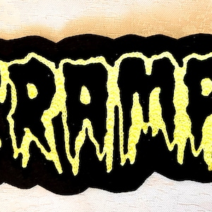 Peut inclure: Un écusson noir avec le nom du groupe "THE CRAMPS" brodé en jaune. La typographie est stylisée avec un effet dégoulinant. Le patch a une bordure noire et est destiné aux vêtements ou accessoires.