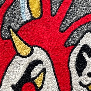 Puede incluir: Un parche textil que presenta una forma central de color rojo vibrante con l&iacute;neas radiantes, acentuada por una protuberancia amarilla en forma de cuerno, &aacute;reas blancas con contornos negros y detalles adicionales en amarillo y azul claro sobre un fondo gris.