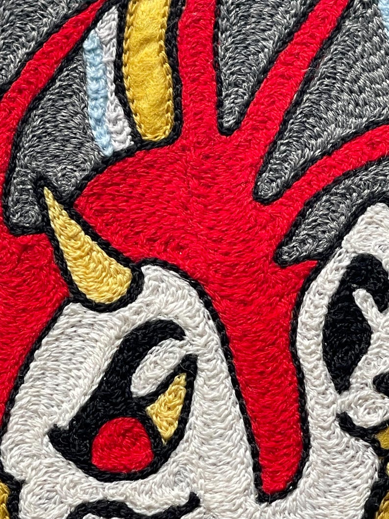 Puede incluir: Un parche textil que presenta una forma central de color rojo vibrante con l&iacute;neas radiantes, acentuada por una protuberancia amarilla en forma de cuerno, &aacute;reas blancas con contornos negros y detalles adicionales en amarillo y azul claro sobre un fondo gris.