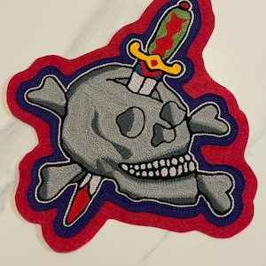 Könnte beinhalten: Gestickter Aufnäher mit einem grauen Totenkopf mit gekreuzten Knochen, durchbohrt von einem Dolch. Der Aufnäher hat einen roten Rand mit einer blauen und weißen Kontur. Der Dolch hat einen gelben Griff und eine rote und grüne Klinge.