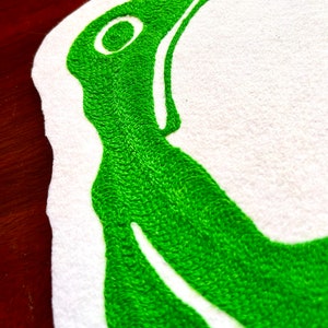 K&ouml;nnte beinhalten: Gr&uuml;ne Stickerei auf wei&szlig;em Stoffhintergrund. Das Design ist ein stilisierter Buchstabe "C" mit einem kleinen Kreis innerhalb der Kurve.