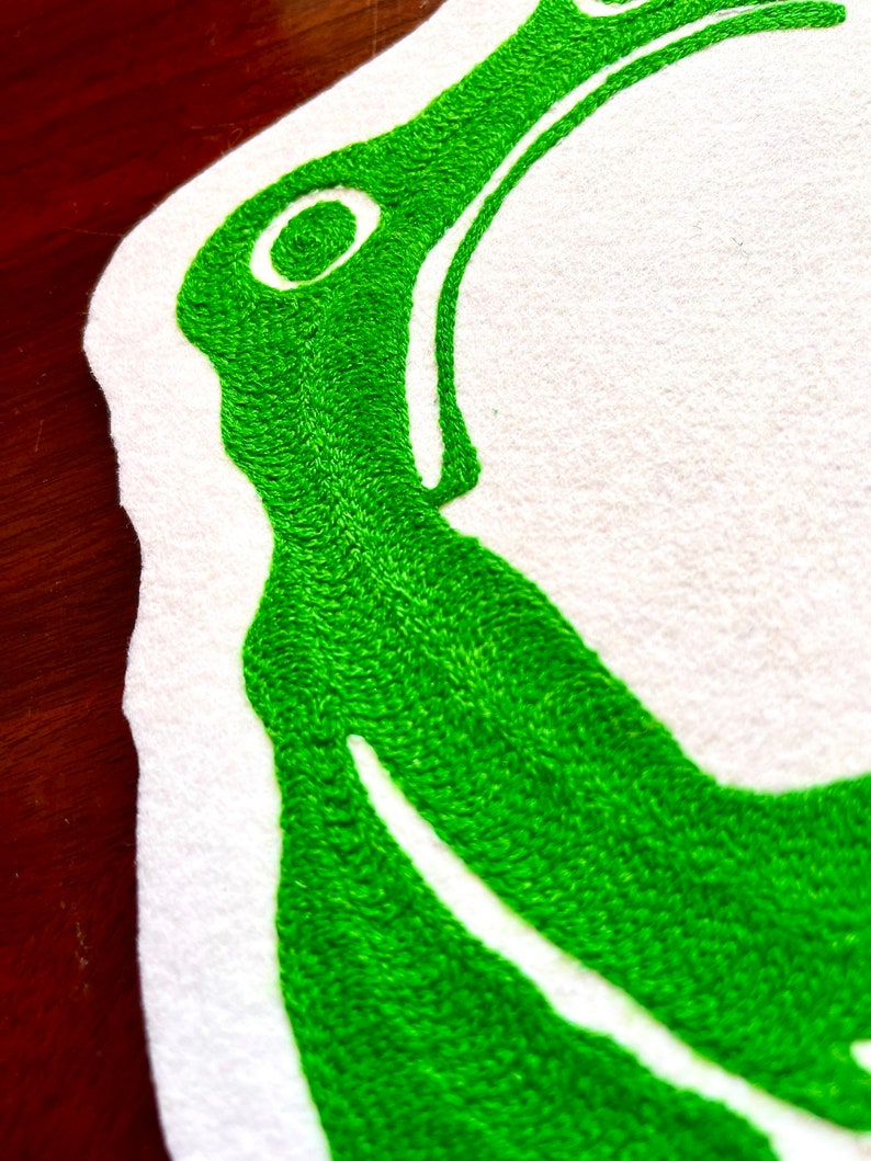 K&ouml;nnte beinhalten: Gr&uuml;ne Stickerei auf wei&szlig;em Stoffhintergrund. Das Design ist ein stilisierter Buchstabe "C" mit einem kleinen Kreis innerhalb der Kurve.