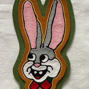 May include: A green, orange, and yellow felt patch with an embroidered image of Bugs Bunny. The patch features a white outline and a red bow tie.