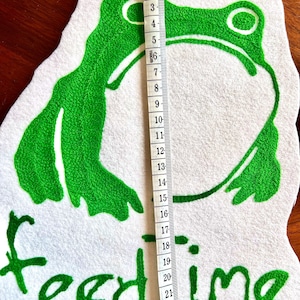 K&ouml;nnte beinhalten: Gr&uuml;ne gestickte Frosch-Applikation auf wei&szlig;em Filzgrund mit dem Text "feed time" in gr&uuml;ner Stickerei. Die Applikation ist etwa 15 cm hoch.