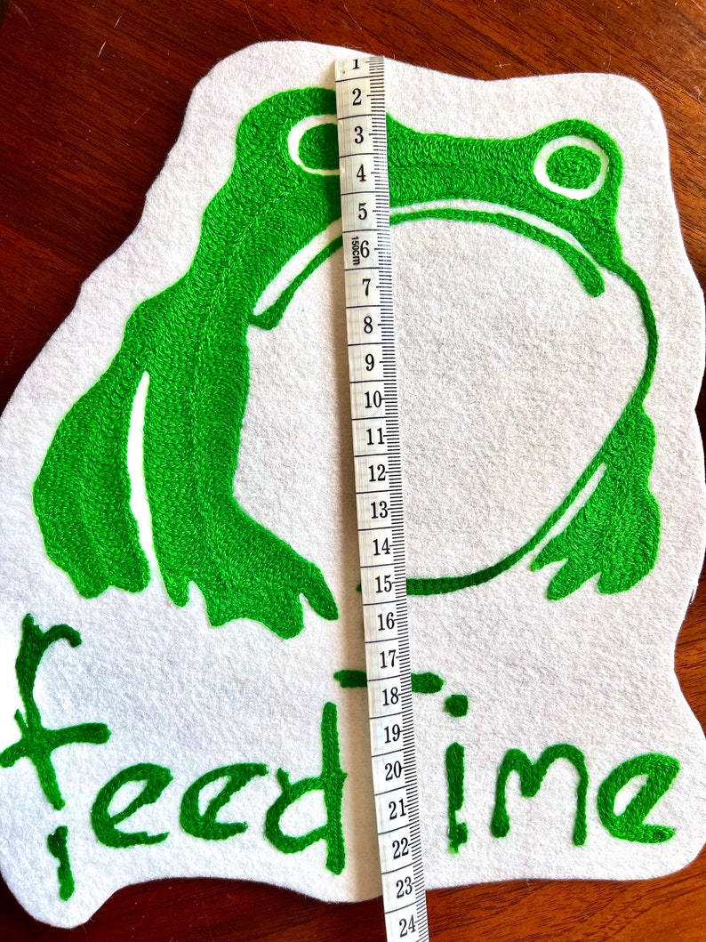 K&ouml;nnte beinhalten: Gr&uuml;ne gestickte Frosch-Applikation auf wei&szlig;em Filzgrund mit dem Text "feed time" in gr&uuml;ner Stickerei. Die Applikation ist etwa 15 cm hoch.