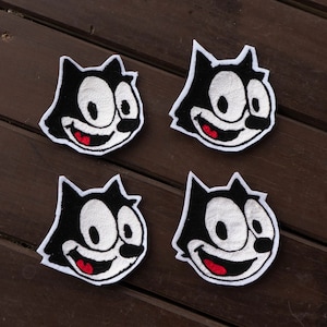 May include: Four black and white embroidered patches of a cartoon cat with a wide grin and large eyes. The patches are on a wooden surface.