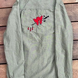 May include: A green long-sleeved button-down shirt with a red heart and dagger graphic on the back. The graphic features two hearts pierced by a dagger with blood dripping from the wound.