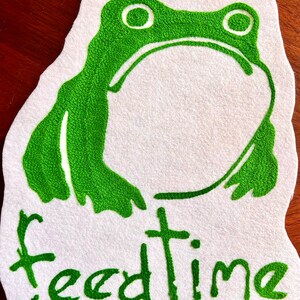 K&ouml;nnte beinhalten: Gr&uuml;ne gestickte Frosch mit gro&szlig;en Augen auf einem wei&szlig;en Filz-Hintergrund. Der Text "feedtime" ist unter dem Frosch gestickt.