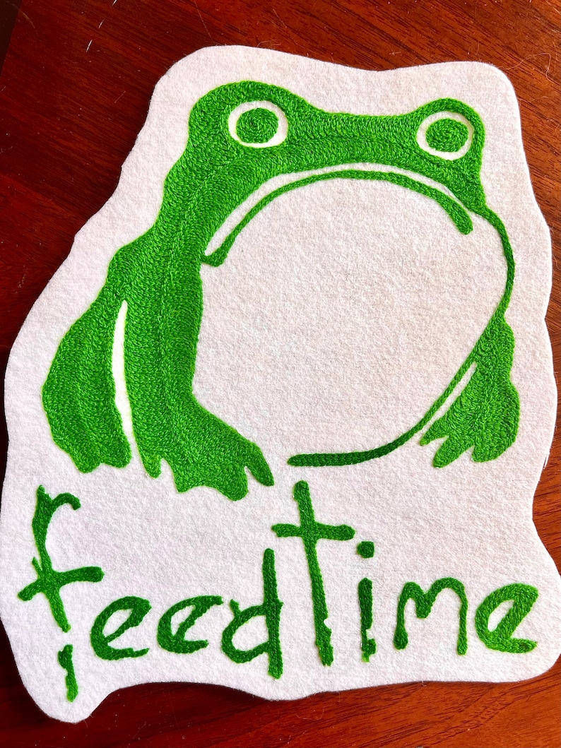 K&ouml;nnte beinhalten: Gr&uuml;ne gestickte Frosch mit gro&szlig;en Augen auf einem wei&szlig;en Filz-Hintergrund. Der Text "feedtime" ist unter dem Frosch gestickt.
