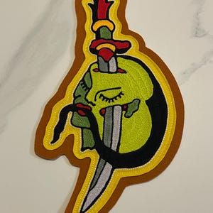 Puede incluir: Parche bordado que presenta una cara verde con los ojos cerrados, un bigote negro y labios rojos, atravesada por una daga. El diseño está delineado en negro, amarillo y marrón, creando un efecto de capas. Para ropa o accesorios.
