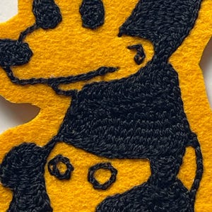 May include: A yellow felt patch featuring black embroidered outlines of two figures. The top figure has a rounded head and dark hair, while the bottom figure wears a dark cap. The embroidery uses thick black thread to define the facial features and hair against the bright yellow felt background.