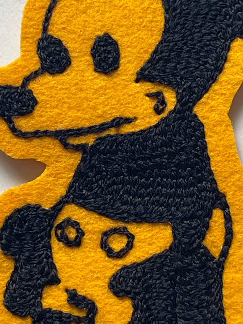 May include: A yellow felt patch featuring black embroidered outlines of two figures. The top figure has a rounded head and dark hair, while the bottom figure wears a dark cap. The embroidery uses thick black thread to define the facial features and hair against the bright yellow felt background.