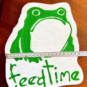 K&ouml;nnte beinhalten: Gr&uuml;ne Filzapplikation eines Frosches mit dem Wort "feedtime" in gr&uuml;n auf einem wei&szlig;en Filzgrund gestickt.