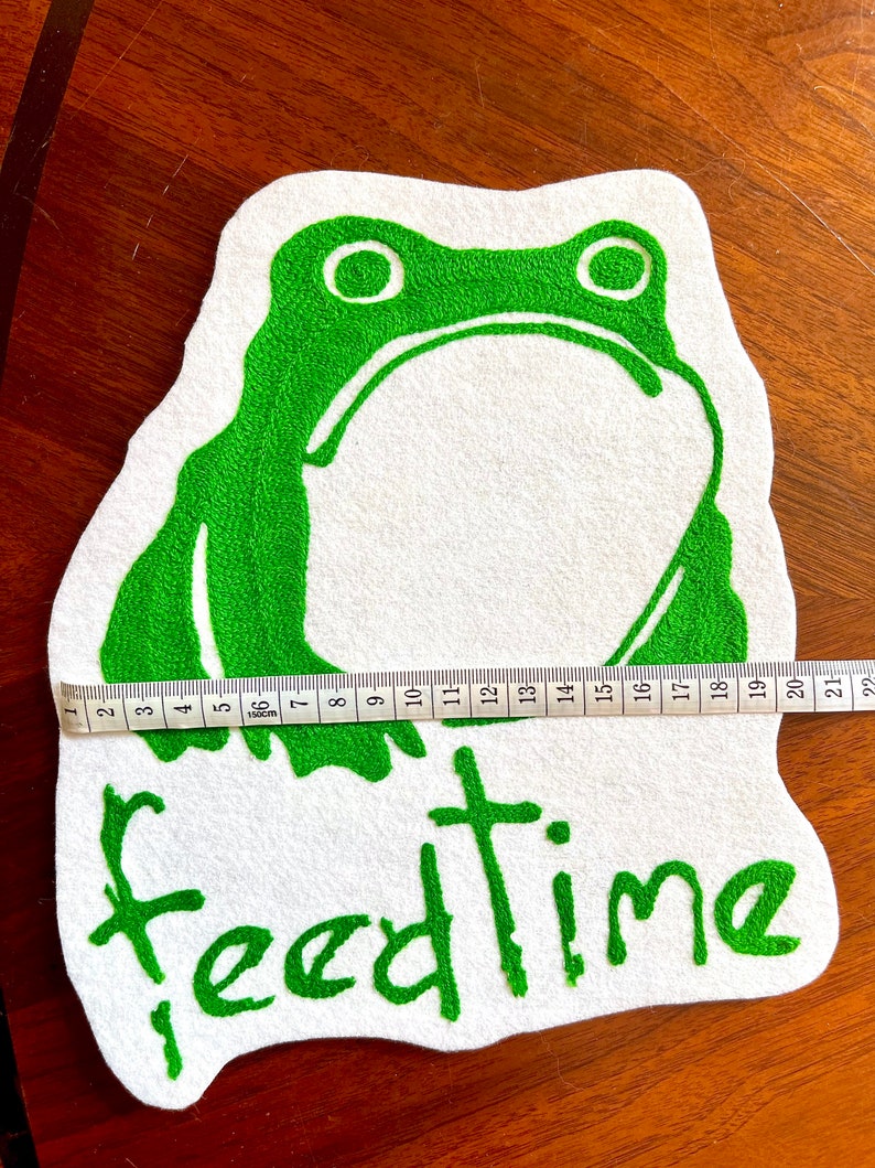 K&ouml;nnte beinhalten: Gr&uuml;ne Filzapplikation eines Frosches mit dem Wort "feedtime" in gr&uuml;n auf einem wei&szlig;en Filzgrund gestickt.