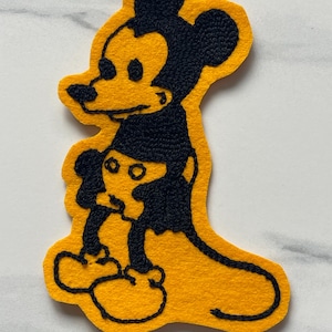 May include: A yellow felt patch featuring a black embroidered outline of a cartoon mouse character. The design shows the character in profile with large ears, a long tail, and rounded feet, rendered in a bold, two-tone color scheme.