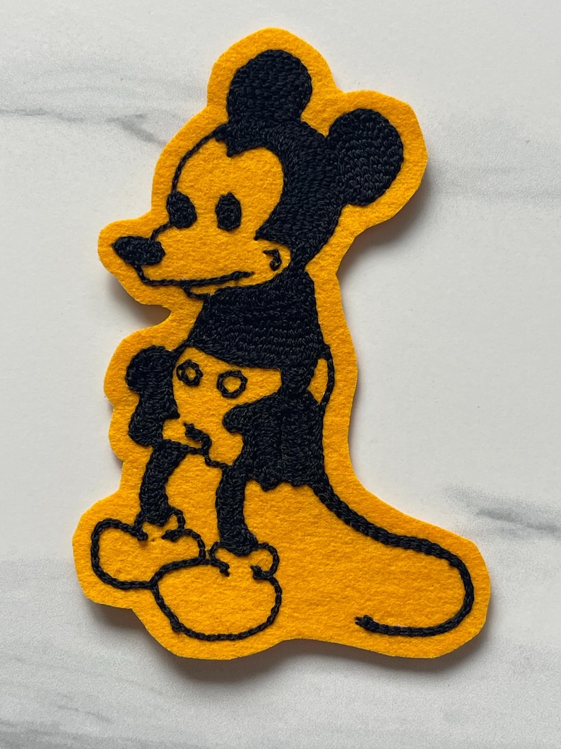 May include: A yellow felt patch featuring a black embroidered outline of a cartoon mouse character. The design shows the character in profile with large ears, a long tail, and rounded feet, rendered in a bold, two-tone color scheme.