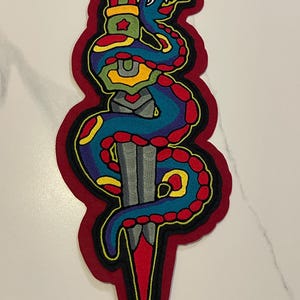 Peut inclure: Un écusson brodé coloré représentant un serpent bleu enroulé autour d'un poignard gris. L'écusson a une bordure rouge avec des détails noirs et jaunes. Le serpent a des taches rouges et une langue jaune.