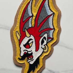 Puede incluir: Un parche bordado que muestra la cabeza de una criatura estilizada con piel blanca, una cresta roja y detalles en negro. El diseño incluye un borde amarillo sobre una base de fieltro color canela. La criatura tiene un pequeño cuerno amarillo y un pendiente dorado.