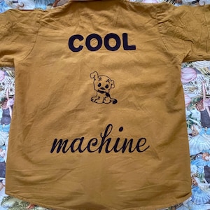 May include: Mustard yellow button-up shirt with short sleeves. The back of the shirt features the word "COOL" above a cartoon dog illustration, and the word "machine" below.