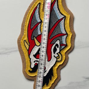 Puede incluir: Un parche de tela con un gr&aacute;fico bordado de una criatura estilizada con detalles en rojo, blanco, negro y gris sobre un fondo de fieltro color canela. Una cinta m&eacute;trica blanca se coloca verticalmente sobre el parche, mostrando 26,5 cm de largo.