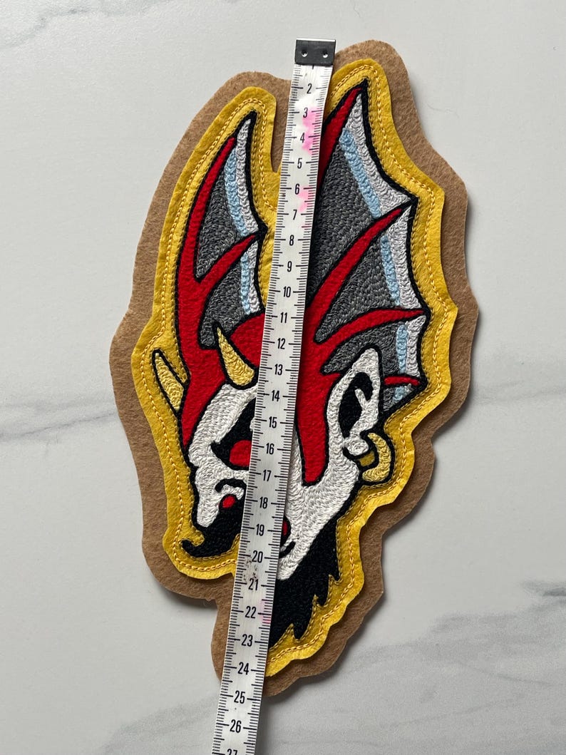 Puede incluir: Un parche de tela con un gr&aacute;fico bordado de una criatura estilizada con detalles en rojo, blanco, negro y gris sobre un fondo de fieltro color canela. Una cinta m&eacute;trica blanca se coloca verticalmente sobre el parche, mostrando 26,5 cm de largo.