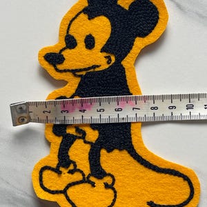 May include: A yellow felt patch featuring a black embroidered outline of a cartoon mouse character. A white measuring tape is placed across the center of the patch, showing a width of approximately 4.5 inches from the edge of the character's body.