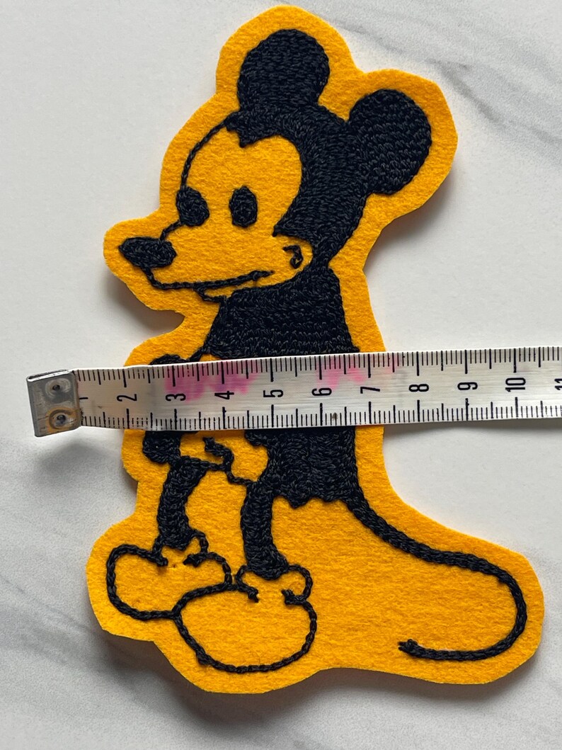 May include: A yellow felt patch featuring a black embroidered outline of a cartoon mouse character. A white measuring tape is placed across the center of the patch, showing a width of approximately 4.5 inches from the edge of the character's body.