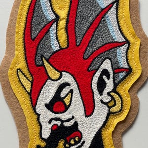 Puede incluir: Un parche de tela con un dise&ntilde;o bordado de una cabeza estilizada con una cresta roja brillante, piel blanca y detalles negros. La figura tiene un cuerno amarillo, un pendiente de aro dorado y est&aacute; delineada con hilo amarillo sobre fieltro beige.