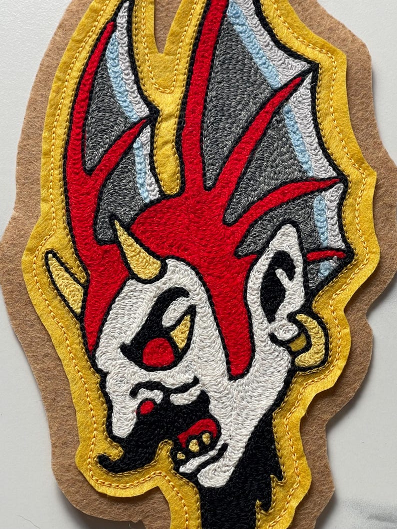 Puede incluir: Un parche de tela con un dise&ntilde;o bordado de una cabeza estilizada con una cresta roja brillante, piel blanca y detalles negros. La figura tiene un cuerno amarillo, un pendiente de aro dorado y est&aacute; delineada con hilo amarillo sobre fieltro beige.
