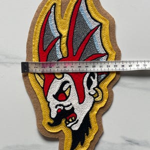 Puede incluir: Un parche bordado que presenta una cabeza de personaje estilizada con detalles en rojo, blanco, negro y gris sobre un fondo de fieltro color canela. Una cinta m&eacute;trica sobre el parche indica un ancho de unos 14 cm en su parte m&aacute;s ancha.