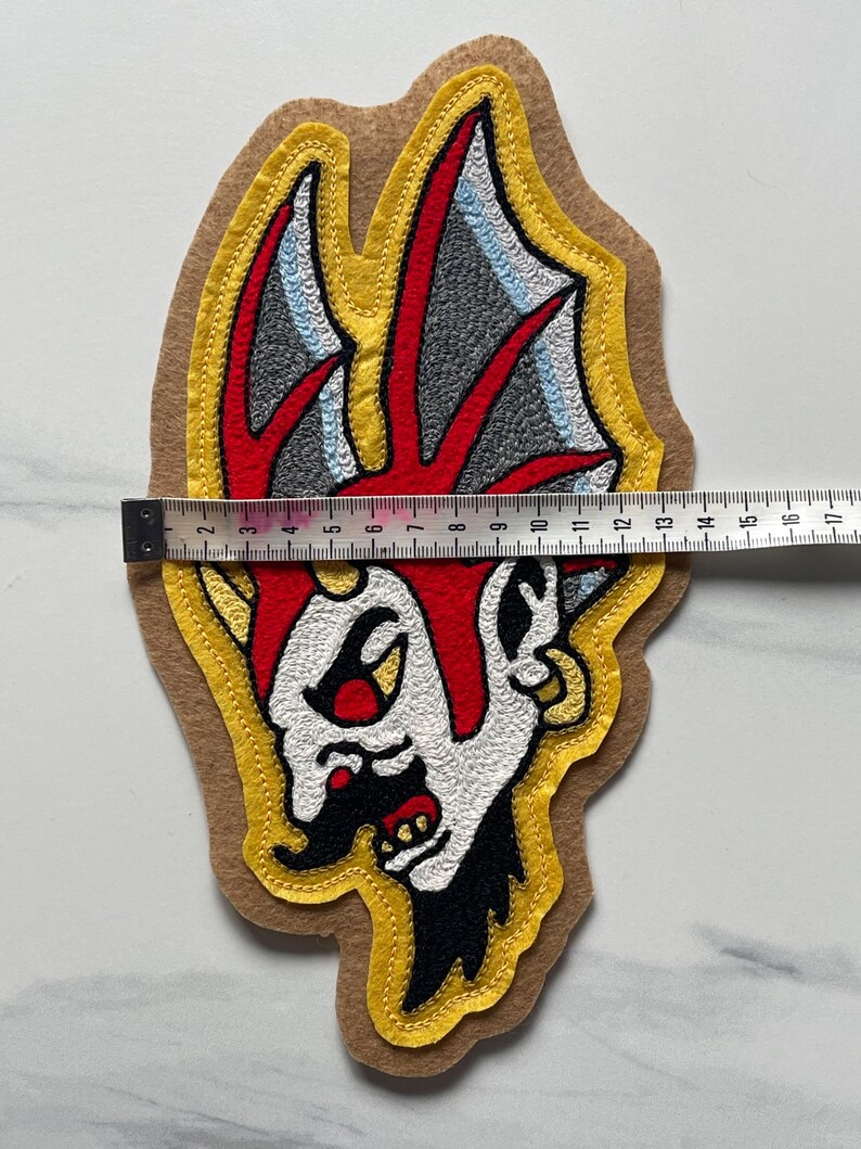 Puede incluir: Un parche bordado que presenta una cabeza de personaje estilizada con detalles en rojo, blanco, negro y gris sobre un fondo de fieltro color canela. Una cinta m&eacute;trica sobre el parche indica un ancho de unos 14 cm en su parte m&aacute;s ancha.