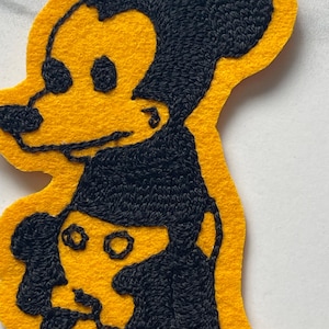 May include: A yellow felt patch featuring black embroidered outlines of a cartoon mouse character. The design shows a larger mouse head positioned above a smaller mouse figure, with detailed black stitching creating the facial features and ears.