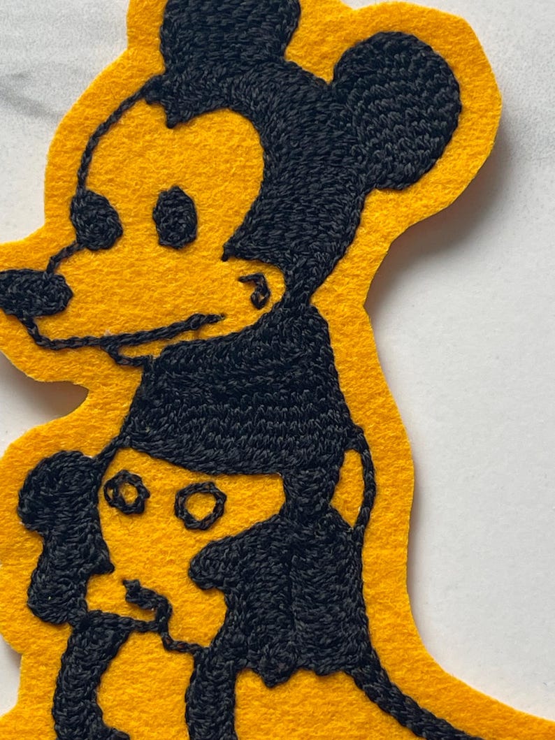 May include: A yellow felt patch featuring black embroidered outlines of a cartoon mouse character. The design shows a larger mouse head positioned above a smaller mouse figure, with detailed black stitching creating the facial features and ears.