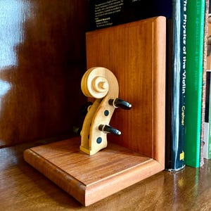 May include: A wooden bookend featuring a violin scroll design. The bookend is made of light brown wood and has a rectangular base. The scroll has black tuning pegs. Books with green and black spines are visible in the background.