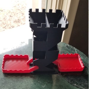 May include: A black and red 3D printed dice tower with a castle design. The tower has three levels and two red trays.