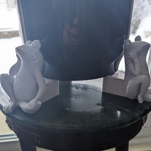 May include: Two white ceramic frog figurines are perched on small white pedestals. The pedestals are positioned on a black and green marble table with a dark wood base.
