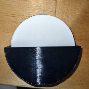 Puede incluir: Un posavasos de plástico negro con un inserto circular blanco. El posavasos está diseñado para sostener un objeto redondo, como una taza o un vaso.