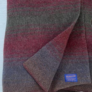 May include: A wool blanket featuring a gradient of horizontal stripes in shades of deep red, olive green, and muted blue. The fabric has a textured, felted appearance. A blue rectangular tag is stitched to the corner with the text Pendleton.