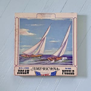 May include: A square jigsaw puzzle box featuring an illustration of two sailboats racing on the ocean under a blue sky. The text reads Americana Jigsaw Puzzle, 700 pieces, and Big 22 x 15 inches. The top includes The America Cup Races text.