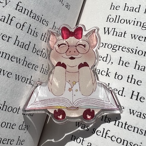 May include: A pink pig wearing glasses and a red bow is sitting on a book with a rainbow design. The pig is holding the book with its paws and has a happy expression on its face.