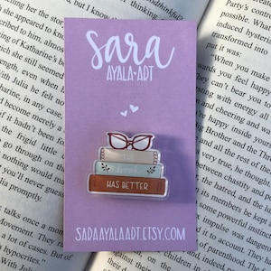 May include: A clear acrylic pin featuring a stack of books with the text "THE BOOK WAS BETTER" and a pair of glasses on top. The pin is on a lavender card with the text "SARA AYALA-ART" and "SARAAYALAART.ETSY.COM".