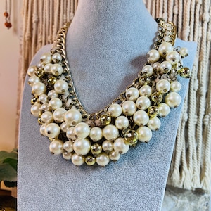 May include: A statement necklace featuring large, creamy white faux pearls and smaller gold-toned beads. The pearls are clustered together, creating a bib-style design. The necklace is attached to a gold-colored chain.