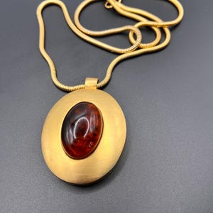 May include: Gold-tone necklace featuring an oval pendant with a brushed finish. The pendant has a central, oval-shaped, dark amber-colored stone. The chain is a thin, flexible, gold-colored snake chain. The jewelry is displayed against a dark background.