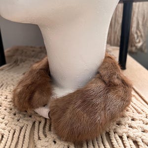 May include: A brown fur collar is displayed on a white mannequin head. The collar is made of soft, textured fur and appears to be a vintage or retro accessory. The collar is designed to be worn around the neck.