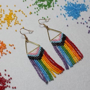 May include: Handmade beaded earrings with a gold-coloured triangular frame and a fringe of colourful beads. The earrings display the colours of the Pride flag and the Transgender Pride flag. Piles of colourful beads are scattered around the earrings.