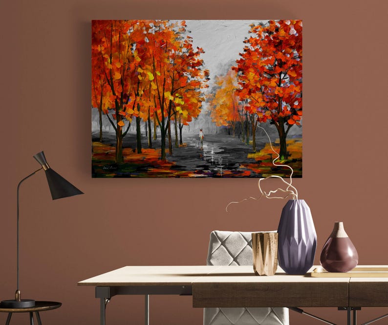 May include: An oil painting depicts a fall scene with trees in vibrant orange and yellow hues lining a path. A figure walks in the distance. The artwork is displayed on a brown wall, with a black lamp and decorative vases on a table in the foreground.