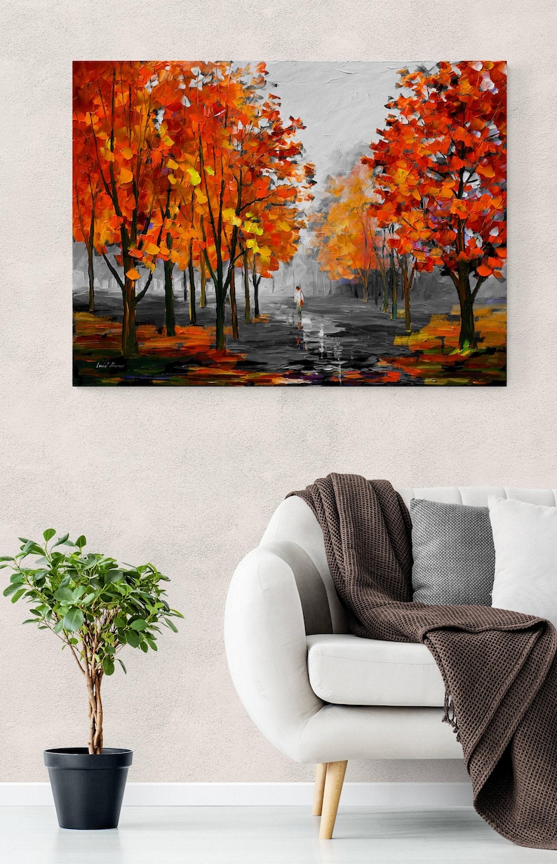 May include: An oil painting depicts a path lined with trees in autumn. The trees have vibrant orange and yellow leaves. A person walks down the path. The painting is displayed on a wall above a white sofa.