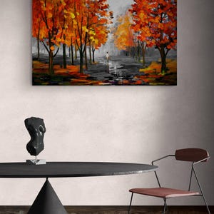 May include: An oil painting depicts a path lined with trees in autumn. The trees have vibrant orange and yellow leaves. A figure walks down the path. The painting hangs above a black table with a sculpture and a chair.